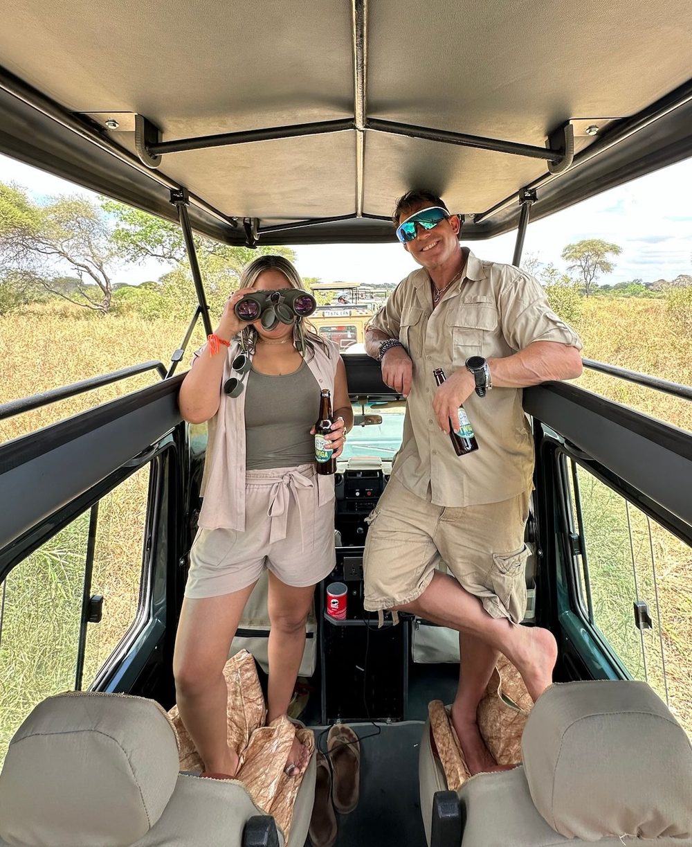 Two travelers in safari clothes standing up through the open roof of a safari vehicle on the savanna, one looking through binoculars